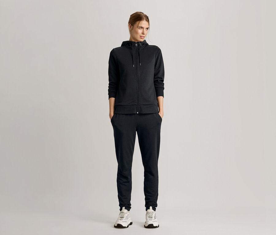 Femme debout, mains dans les poches, portant un survêtement noir avec un sweat à capuche zippé et des baskets.