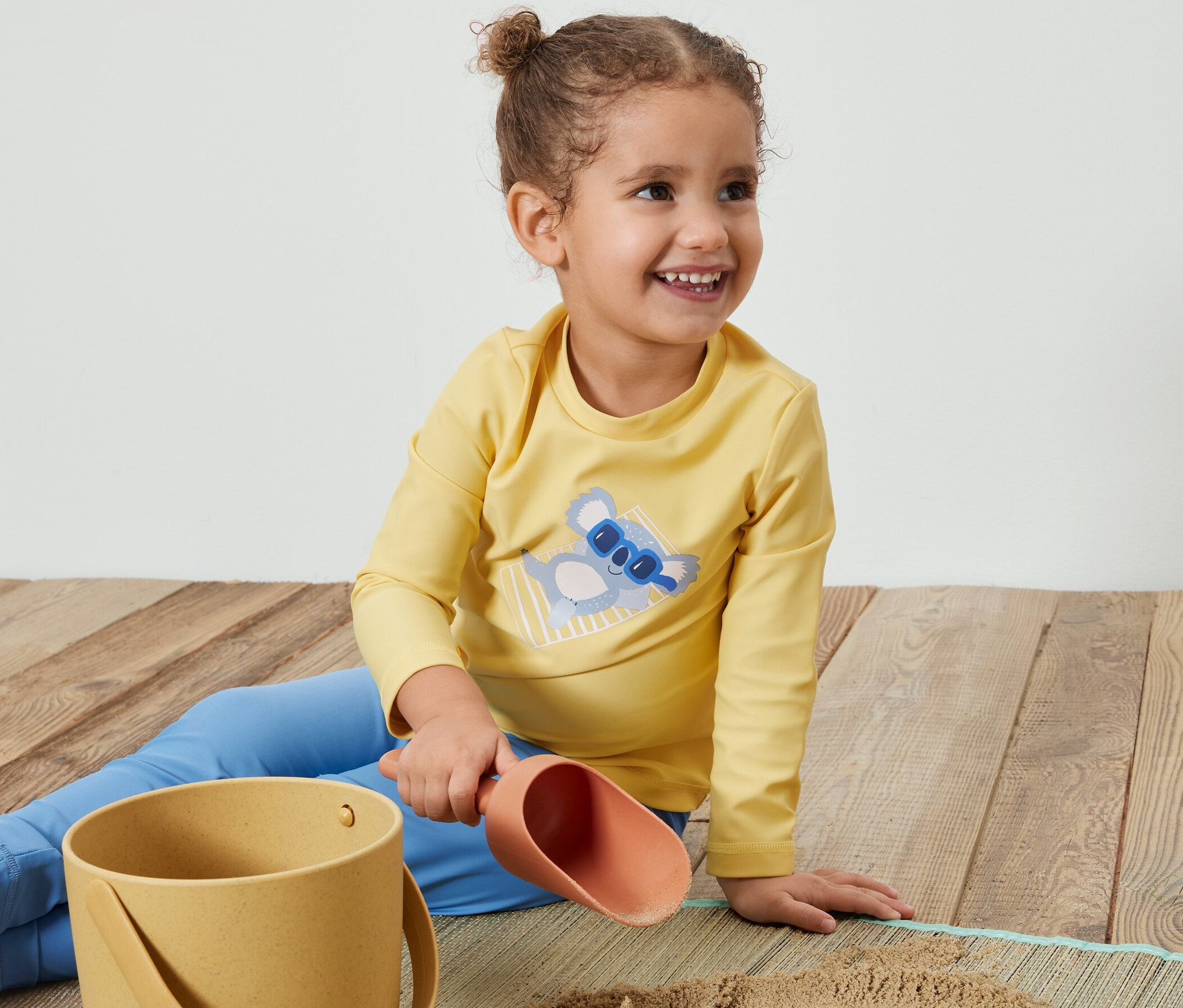 Fillette souriante assise par terre jouant avec du sable et portant un t-shirt jaune pour enfant avec indice de protection UV 80, koala.