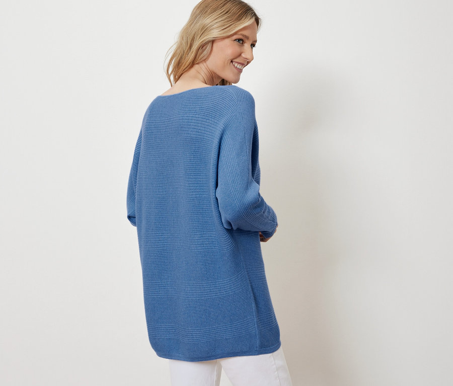 Blonde souriant par-dessus son épaule portant un pull bleu en maille fine et un pantalon blanc.