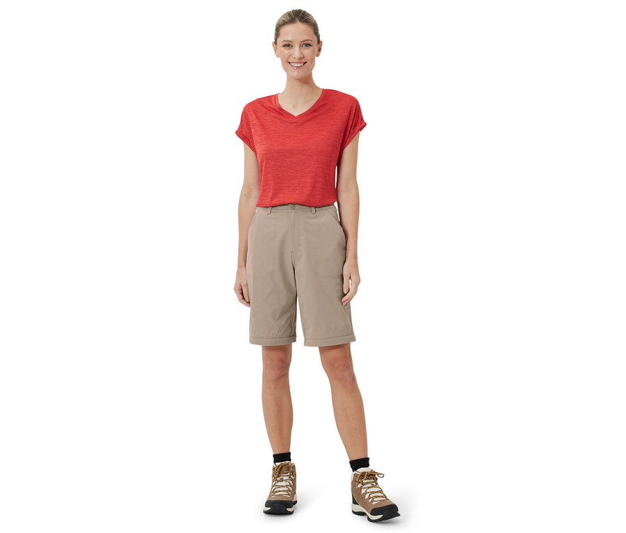 Une femme debout porte un pantalon fonctionnel 2 en 1 beige, un t-shirt rouge et des chaussures marron.
