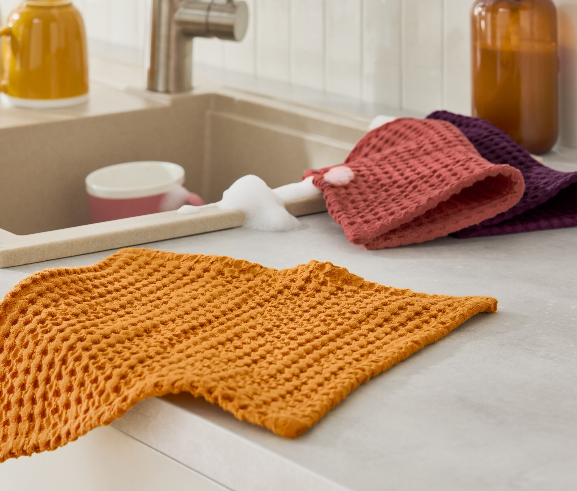 Un torchon orange est posé sur le plan de travail à côté de l'évier avec une éponge et du liquide vaisselle.