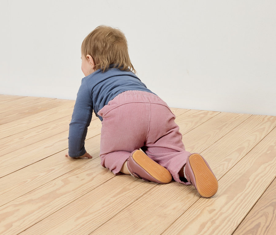 Un bébé rampe sur un plancher en bois, il porte un t-shirt bleu et un pantalon rose.