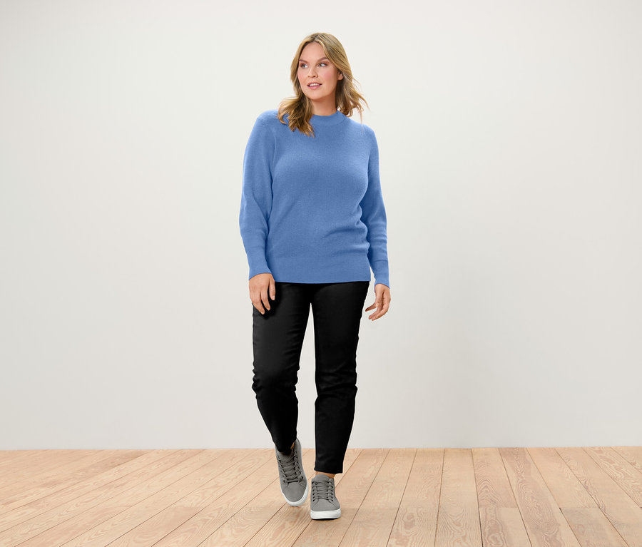 Femme marchant sur un plancher en bois, portant un pull bleu, un pantalon noir et des chaussures grises.