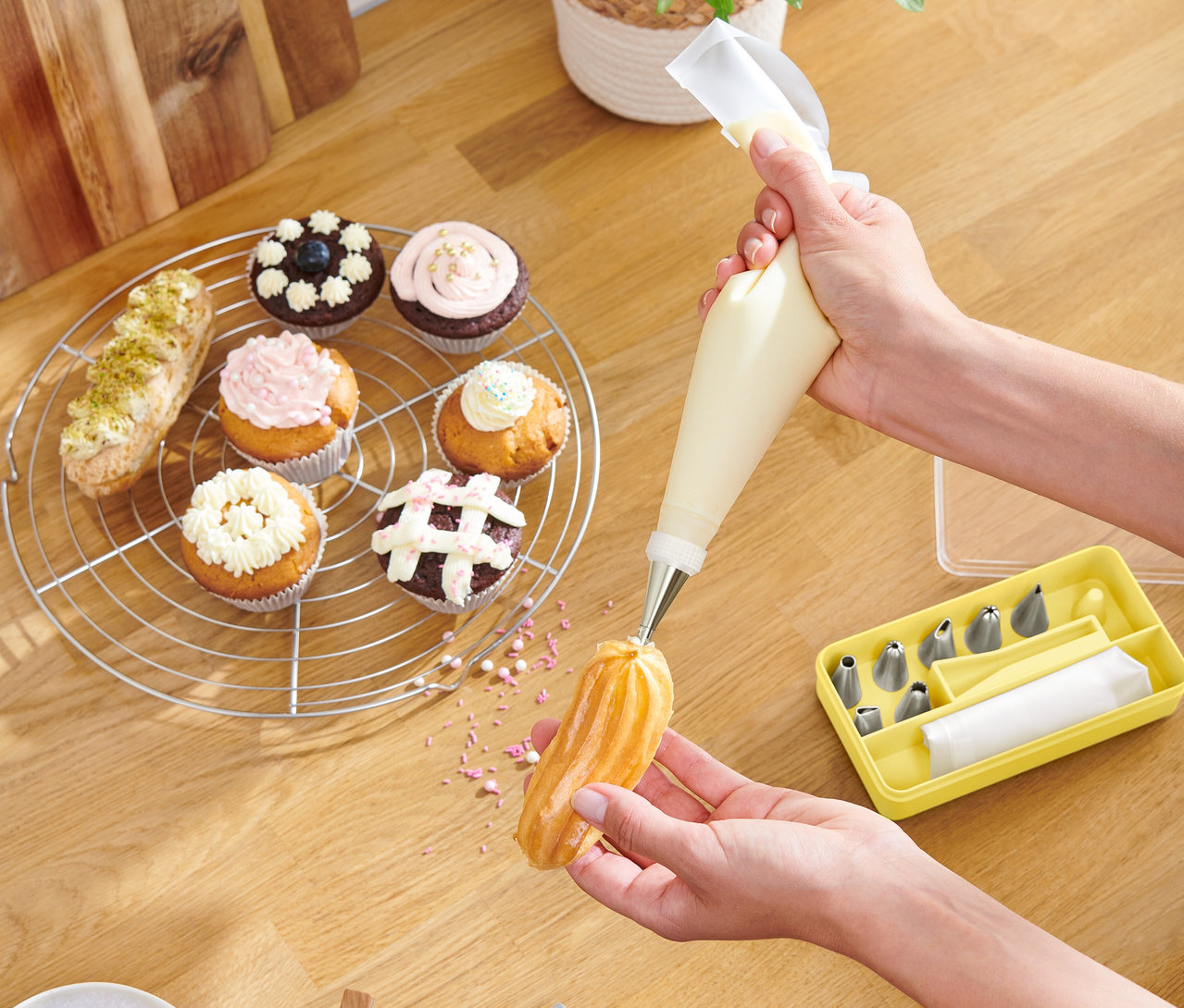 Une personne décore une pâtisserie avec une poche à douille, des cupcakes et une boîte jaune d'embouts sont posés sur une table en bois.