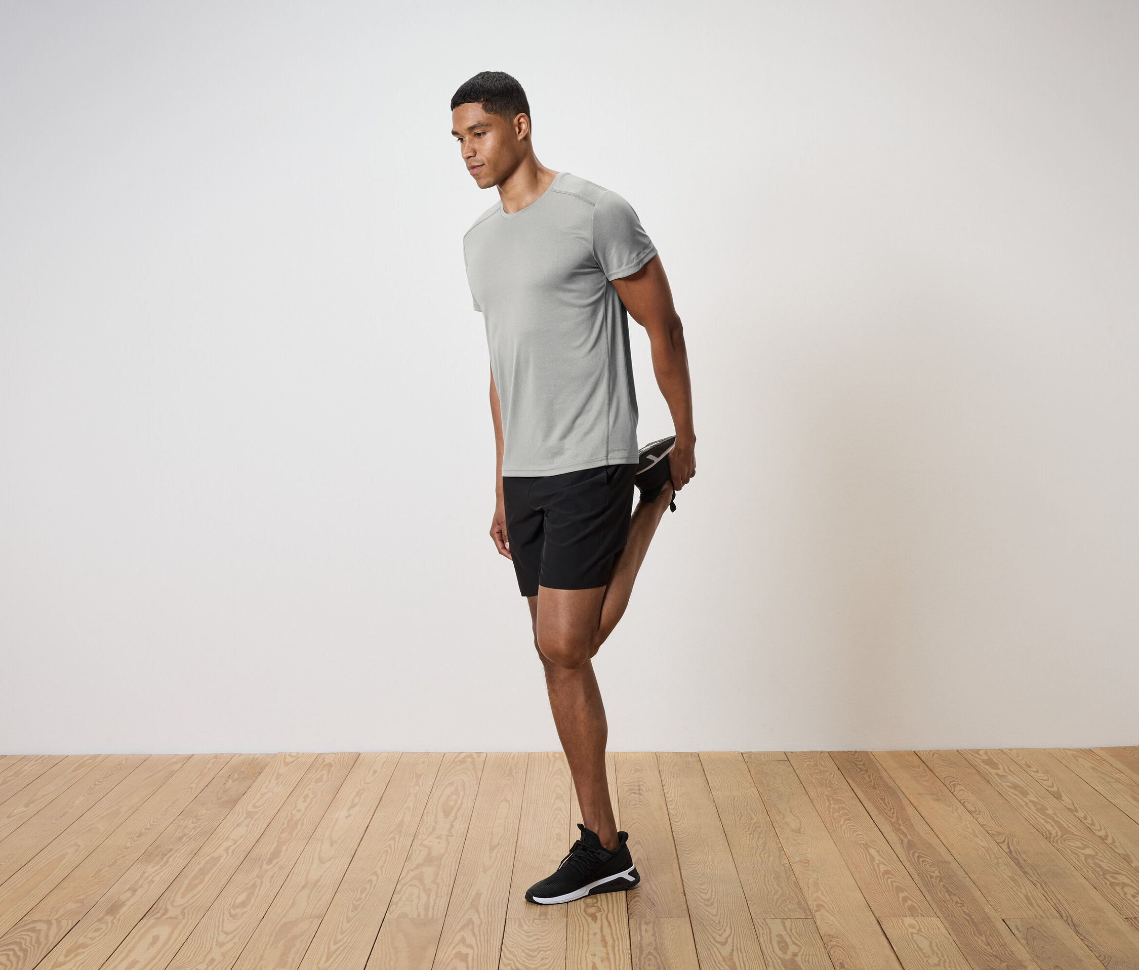 Un homme en t-shirt gris et short noir étire sa jambe sur un plancher en bois.