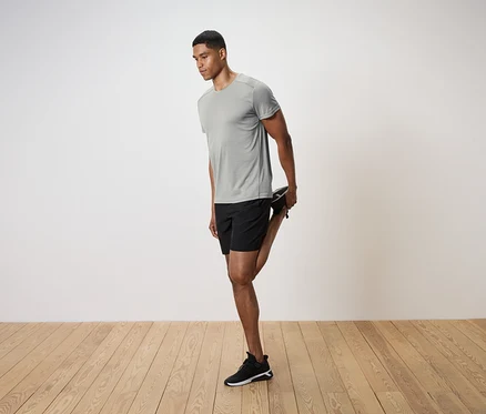 Un homme en t-shirt gris et short noir étire sa jambe sur un plancher en bois.