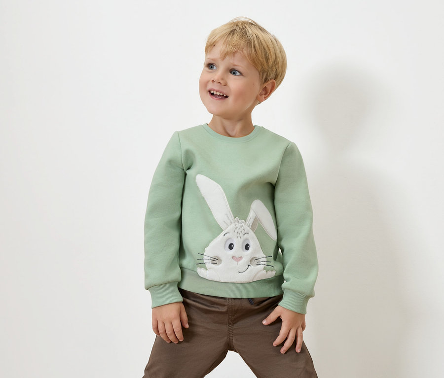 Un garçon portant un pull vert avec un motif de lapin et un pantalon marron se tient devant un fond blanc, regardant vers le haut.