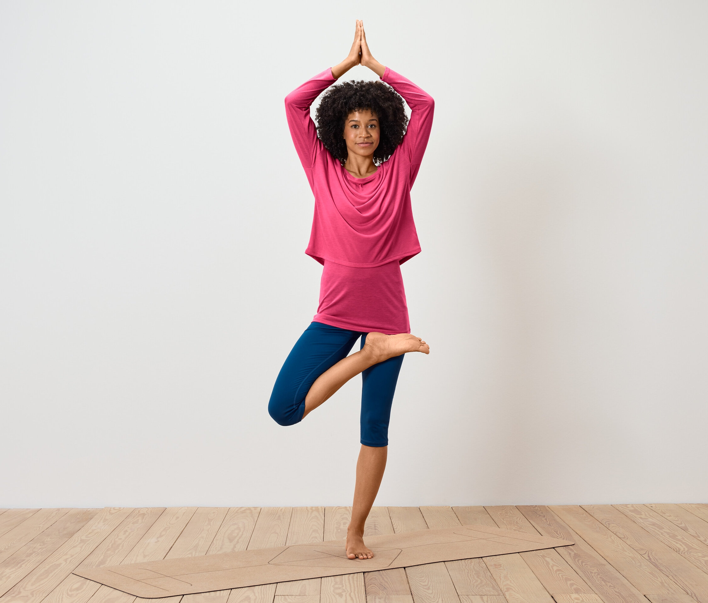 Une femme avec une coiffure afro se tient dans la pose de l'arbre sur un tapis de yoga, portant un haut à manches longues rose et un legging bleu.