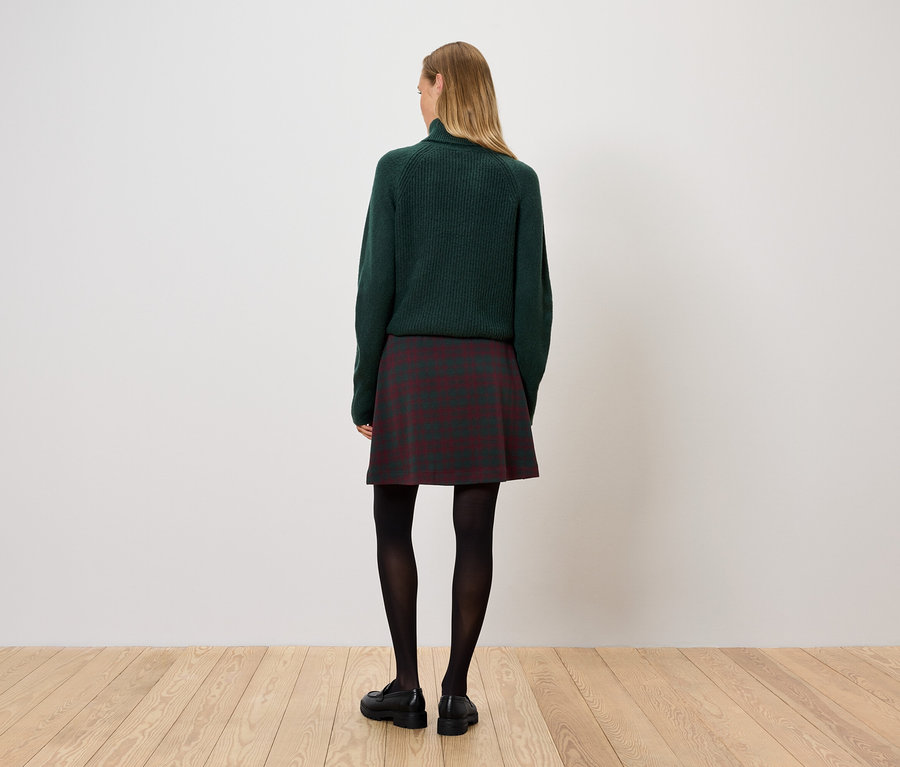 Une femme se tient dos à la caméra, portant un pull vert, une jupe à carreaux et des collants noirs.