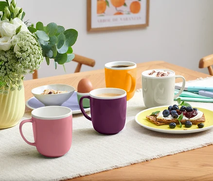 Sur une table, il y a quatre tasses de différentes couleurs, un bol de muesli, un œuf dans un coquetier et une assiette avec une tartine aux myrtilles.