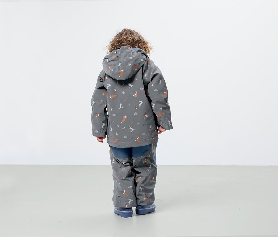 Un garçon aux cheveux bouclés porte une veste et un pantalon gris avec des motifs de renards et d'oiseaux, tourné dos à la caméra.