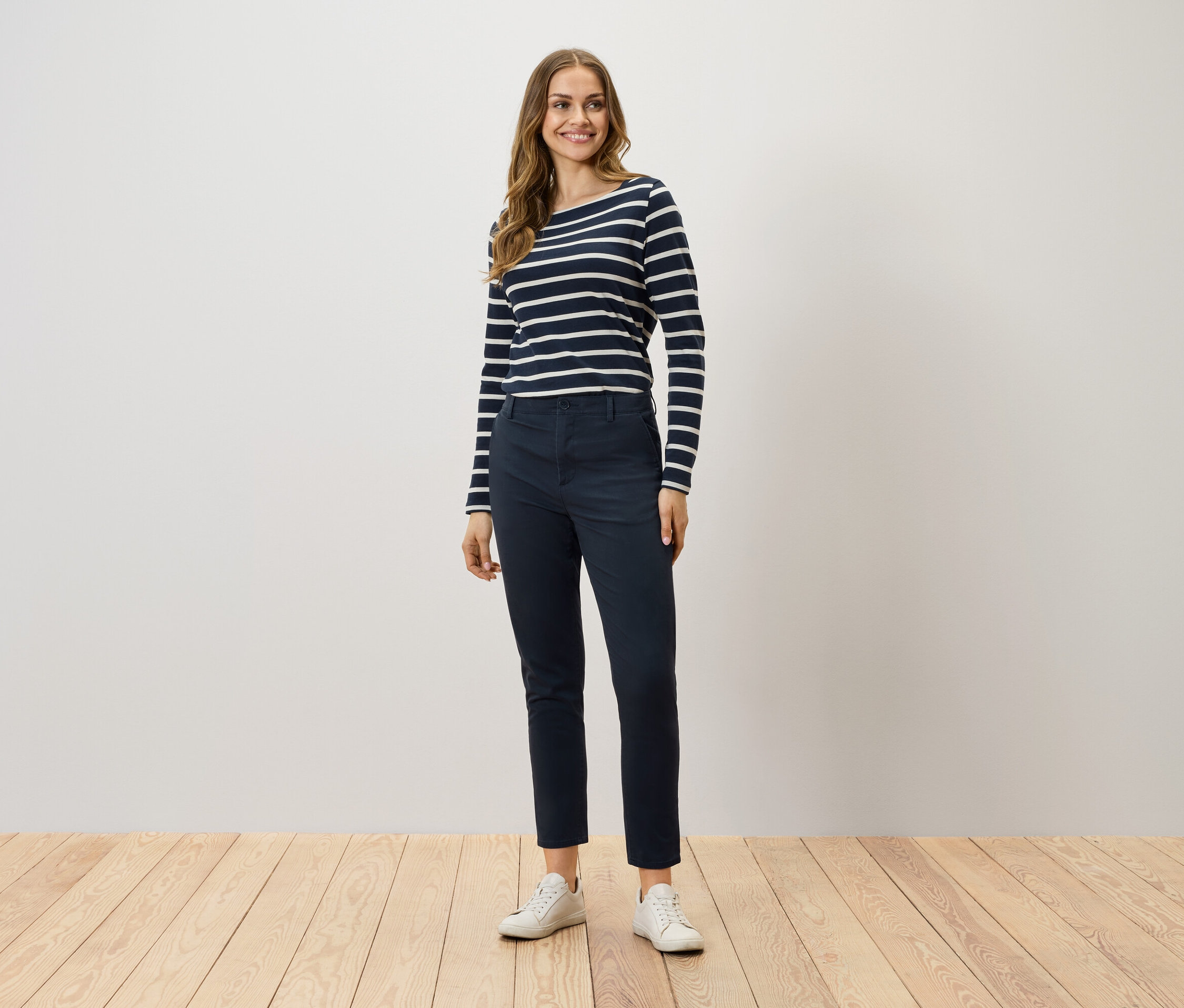 Une femme debout porte un t-shirt rayé à manches longues, un pantalon bleu marine et des baskets blanches.