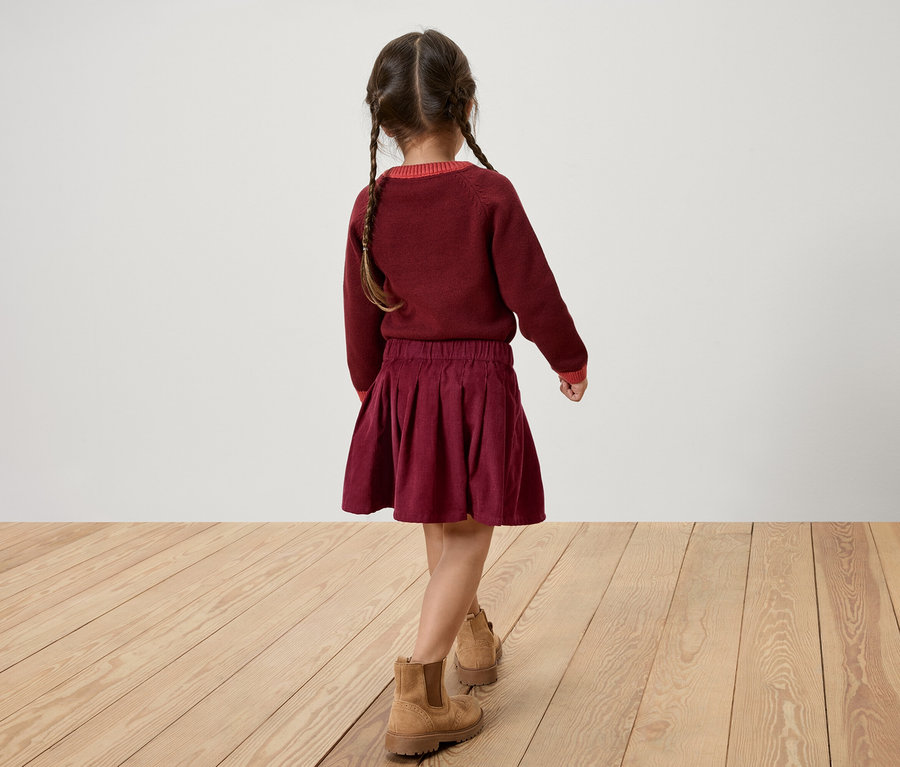 Vue de dos d'une fille portant un pull et une jupe bordeaux, avec des bottes marron, marchant sur un plancher en bois.