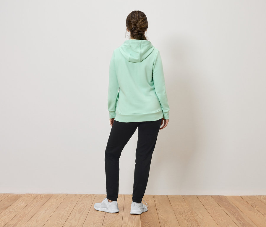 Une femme, vue de dos, porte un sweat à capuche vert clair, un pantalon de jogging noir et des baskets blanches.