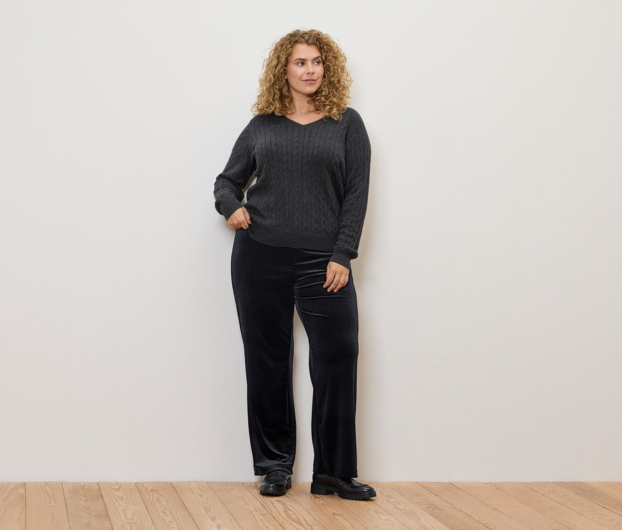 Une femme aux cheveux bouclés pose, portant un pull gris à torsades et un pantalon en velours noir.