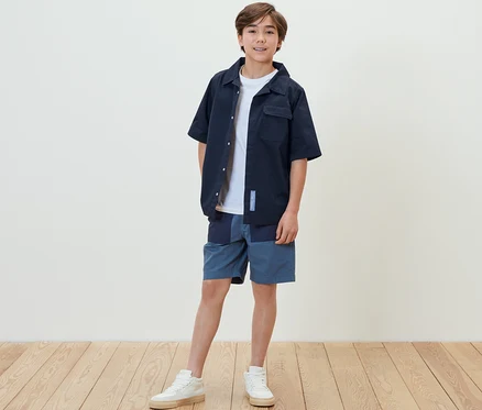 Un garçon debout portant une chemise pour enfant, un short et des baskets.