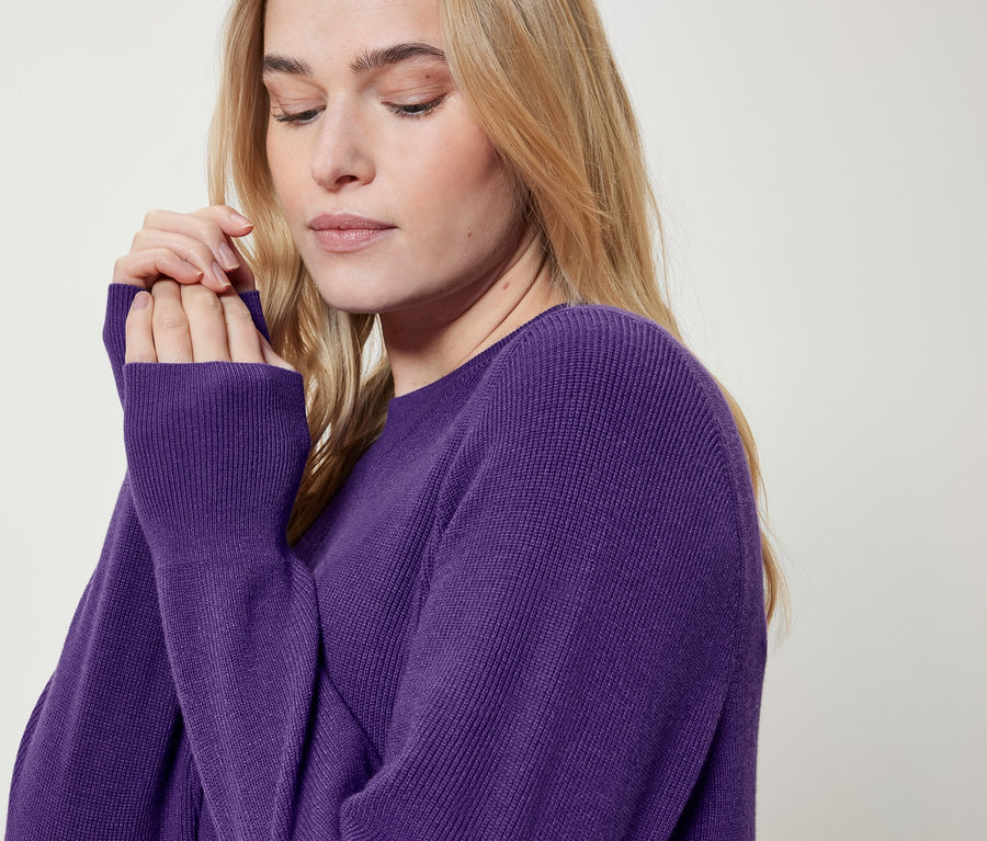 Une femme blonde portant un pull violet porte ses mains à son visage.