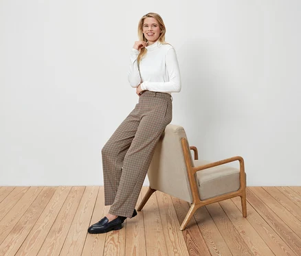 Une femme blonde est assise sur un fauteuil beige, elle porte un col roulé blanc, un pantalon à carreaux marron et des mocassins noirs.