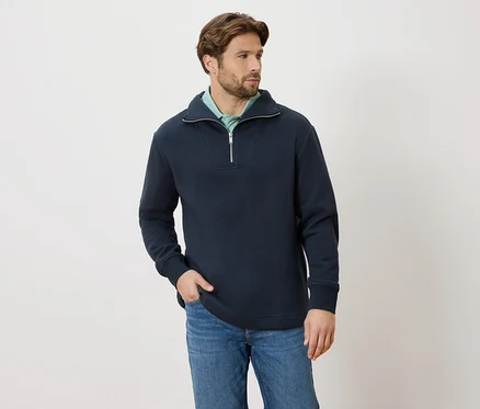 Homme portant un pull bleu marine à fermeture éclair et un jean, la main dans la poche, regardant vers la droite.