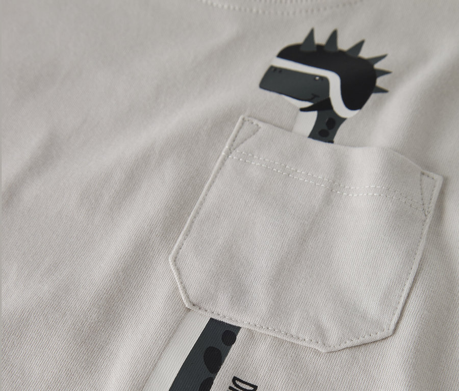Gros plan sur un t-shirt gris clair avec une figure de dinosaure portant un casque imprimée, sortant d'une poche de poitrine. Le t-shirt porte l'inscription "Dino Racer Born to Race".
