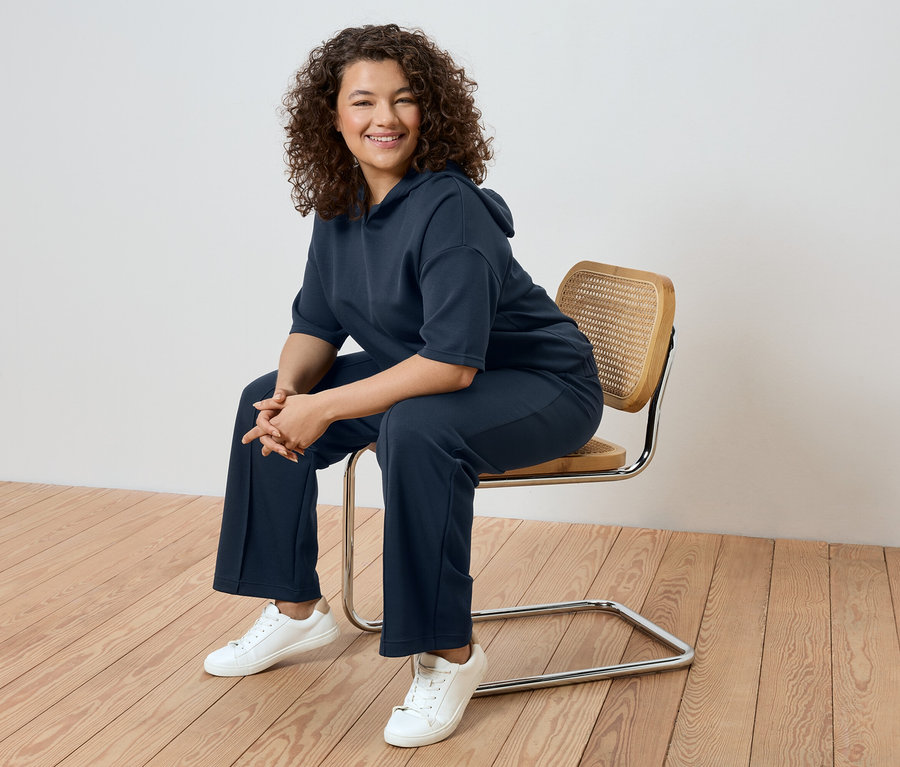 Une femme aux cheveux bouclés est assise sur une chaise, portant un survêtement bleu marine et des baskets blanches.