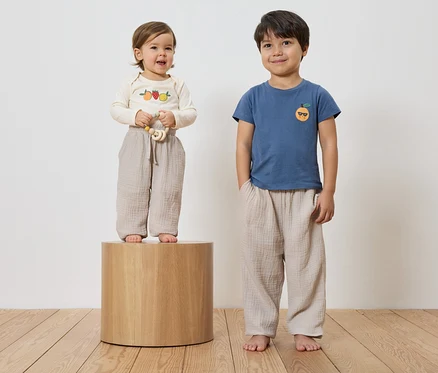 Un bambin avec une chemise brodée et un anneau de dentition se tient sur un socle en bois, à côté d'un petit garçon avec une chemise bleue et une orange brodée, tous deux portant des pantalons amples.
