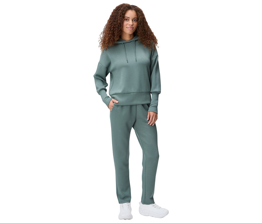 Femme aux cheveux bouclés portant un sweat à capuche vert, un pantalon de jogging et des baskets blanches.