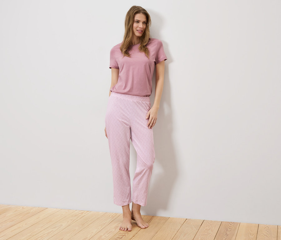 Une femme se tient devant un mur blanc, elle porte un t-shirt rose et un pantalon rose à motifs.