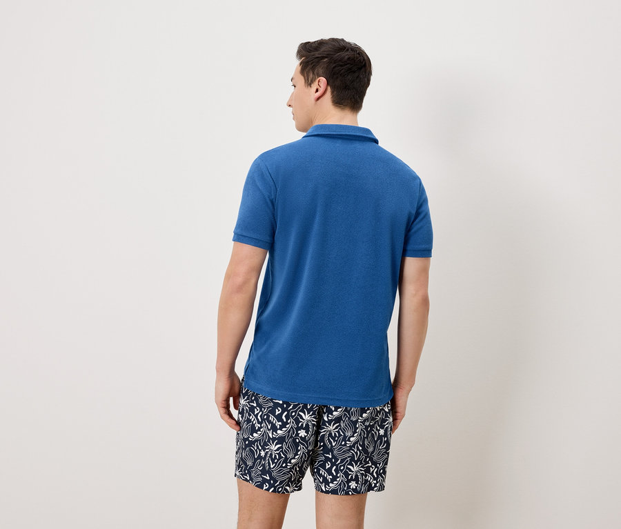 Un homme vu de dos portant un polo bleu et un short de bain bleu et blanc.