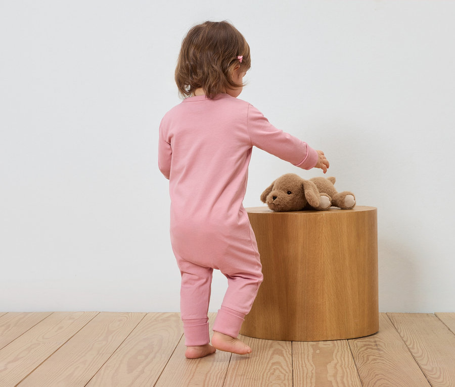 Un enfant en combinaison rose se tient à côté d'un tabouret en bois et touche un chien en peluche.