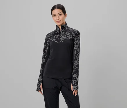 Une femme portant une queue de cheval porte un haut noir à fleurs et un pantalon noir sur un fond gris.