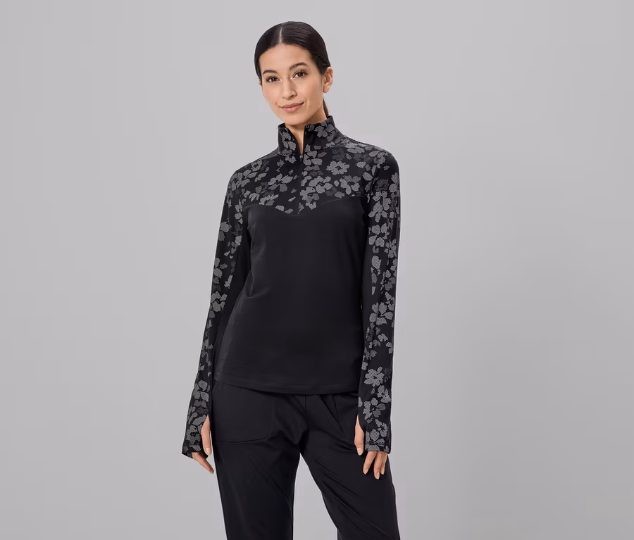 Une femme portant une queue de cheval porte un haut noir à fleurs et un pantalon noir sur un fond gris.