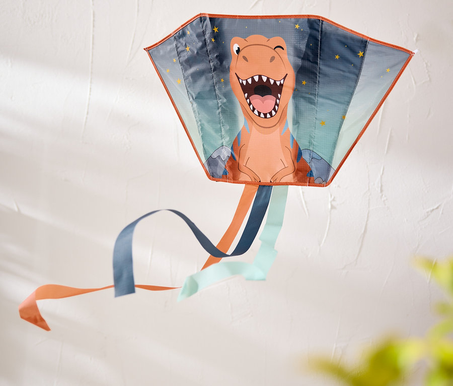 Un cerf-volant coloré avec un visage de dinosaure orange, souriant et clignant de l'œil, et des rubans bleus, oranges et bleu clair, accroché à un mur blanc.