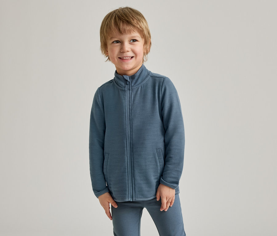 Un jeune garçon aux cheveux blonds sourit, vêtu d'une veste zippée bleue et d'un pantalon assorti, devant un fond clair.
