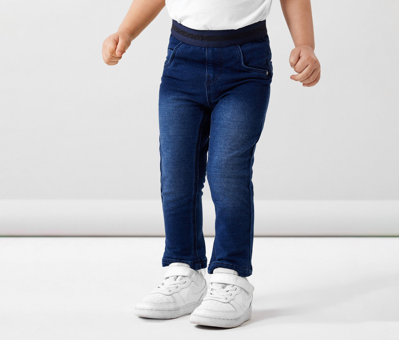 Un enfant porte un jean en sweat NAME IT et des chaussures blanches sur fond blanc.