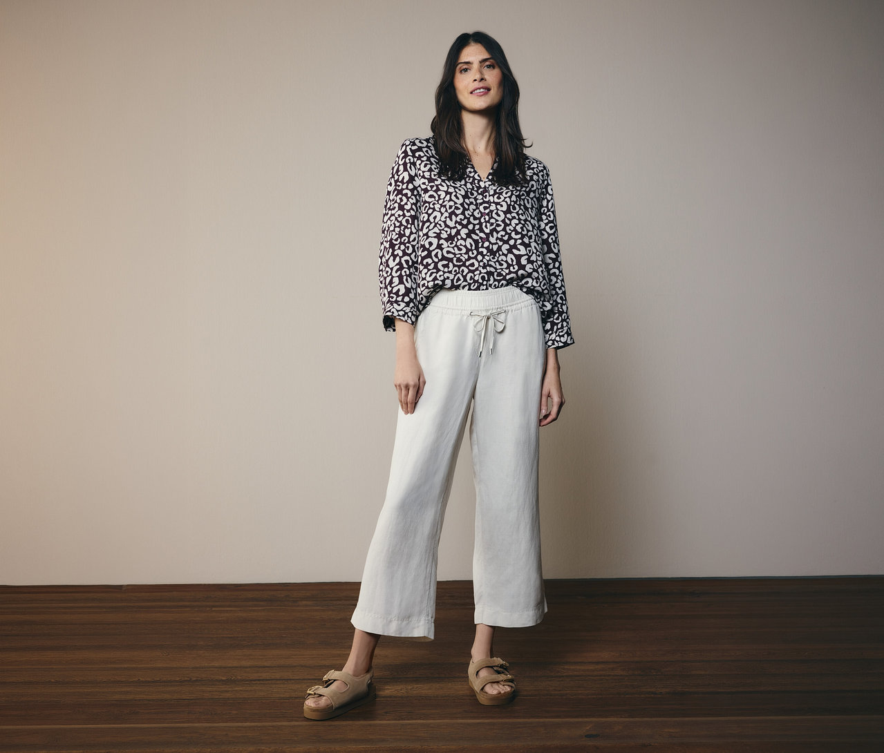 Femme portant une blouse CECIL bordeaux à motif abstrait clair, un pantalon large court beige clair et des sandales beiges, debout devant un mur gris clair.