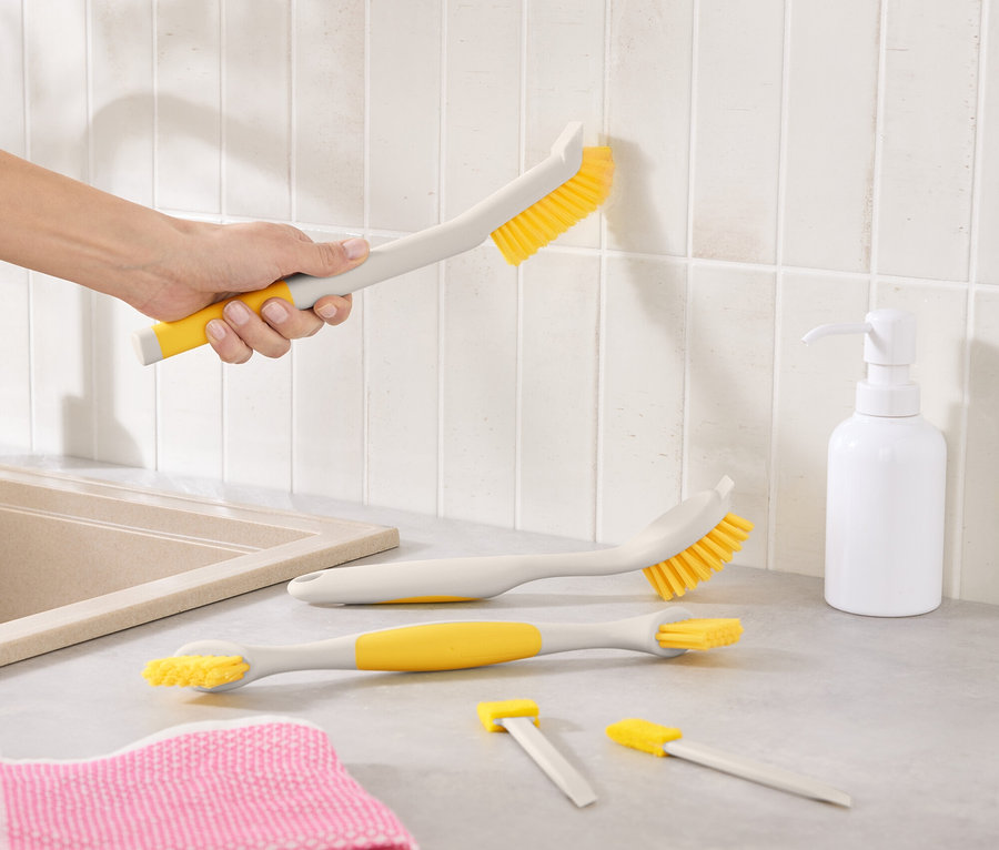 Une main nettoie des carreaux avec une brosse jaune. Sur le comptoir de la cuisine se trouvent d'autres brosses, un tissu rose et un flacon de savon.