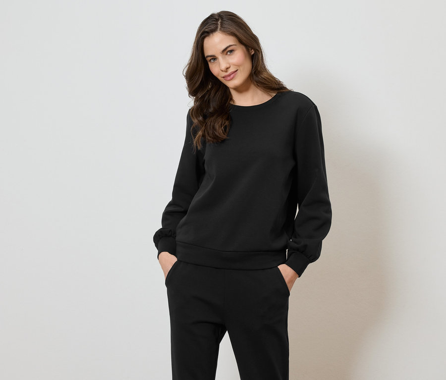 Une femme portant un sweat-shirt et un pantalon noirs pose devant un fond blanc.
