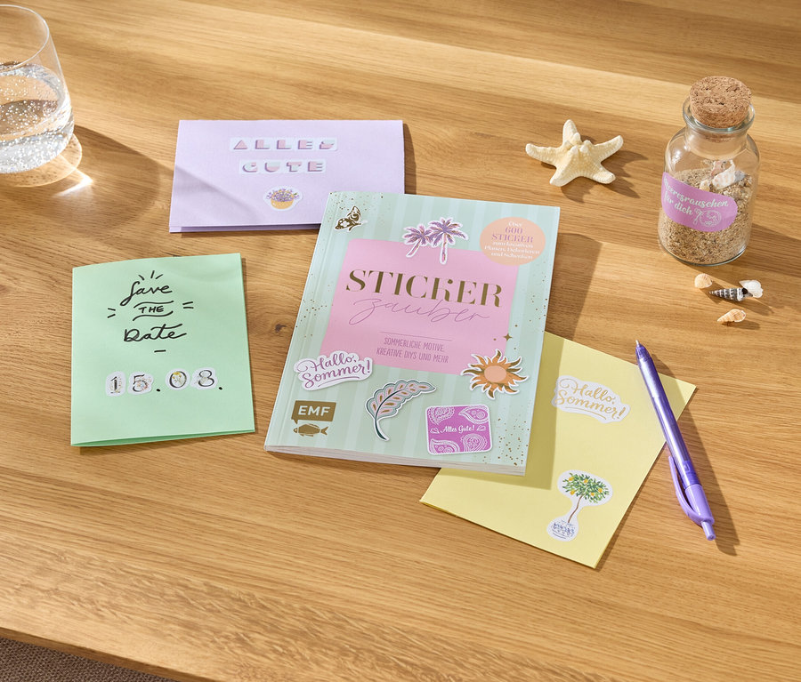 Sur une table, un verre d'eau, des cartes avec des inscriptions, un livre d'autocollants, un stylo, une étoile de mer et une bouteille de sable.