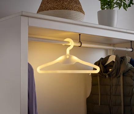 Dans une armoire blanche, un cintre lumineux est suspendu à une barre, à côté d'un gilet et d'une chemise.