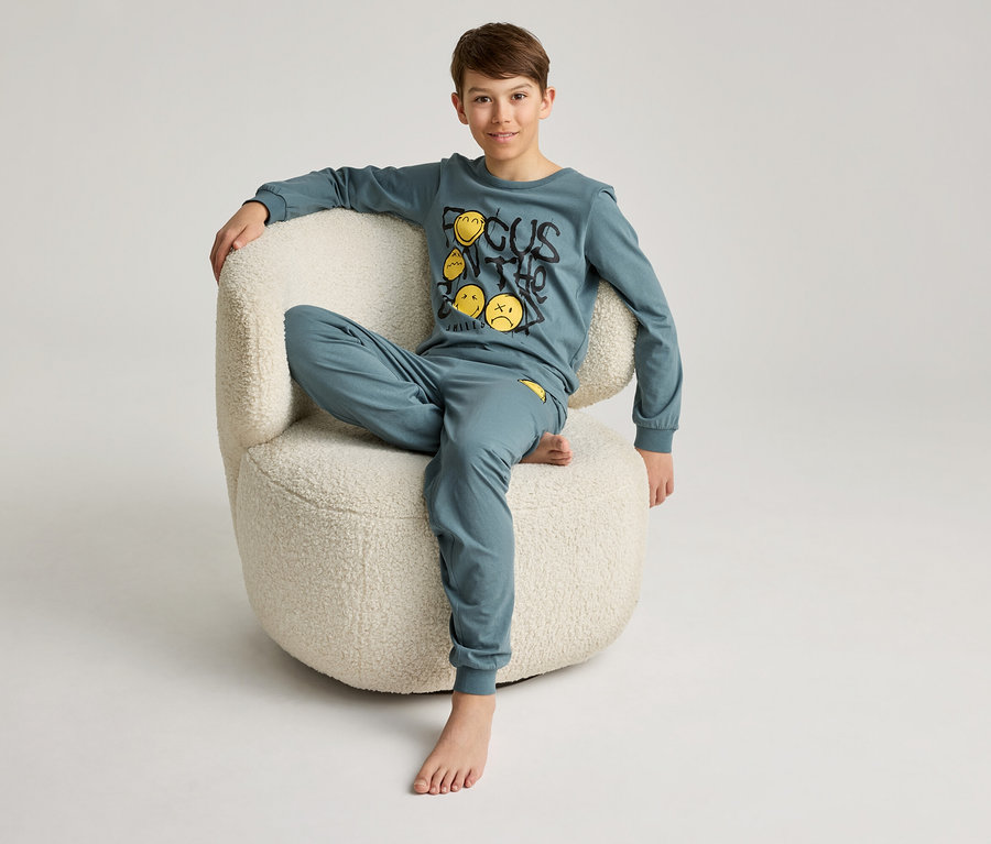 Un garçon est assis pieds nus dans un fauteuil et porte un pyjama bleu pétrole avec un motif de smiley.