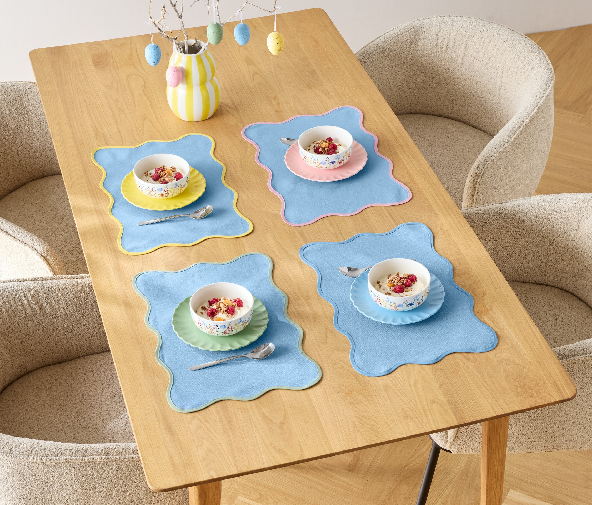 Une table à manger est dressée pour quatre personnes avec des bols de céréales et des sets de table colorés, entourée de quatre chaises.