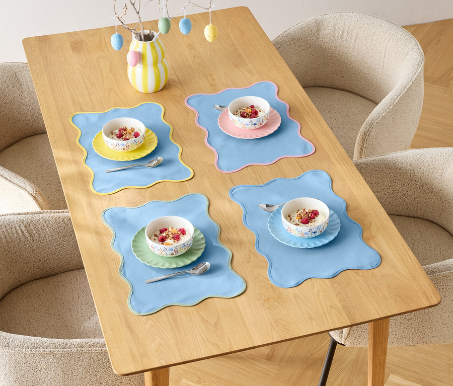 Une table à manger est dressée pour quatre personnes avec des bols de céréales et des sets de table colorés, entourée de quatre chaises.