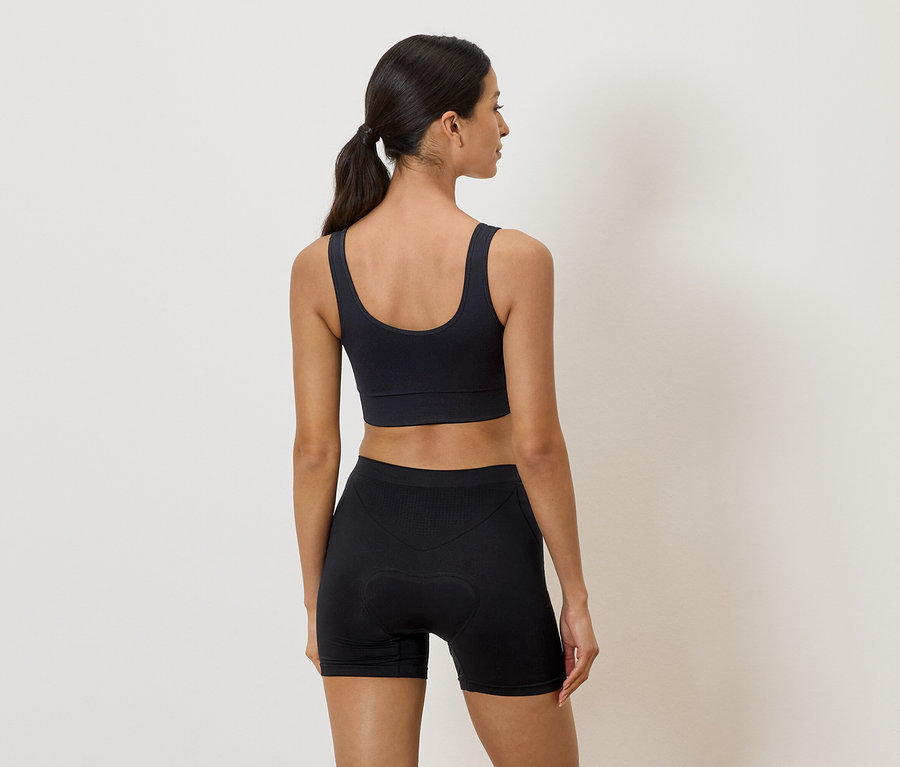 Femme de dos en soutien-gorge de sport noir et short de cycliste sur fond neutre.