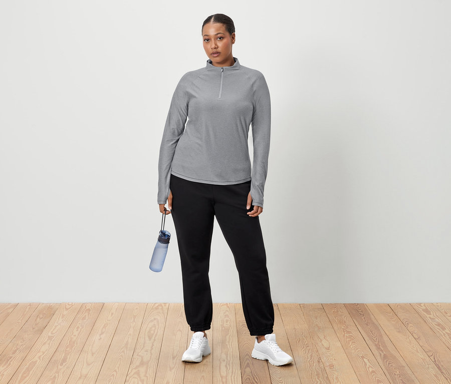 Une femme se tient devant un mur blanc et tient une bouteille d'eau bleue à la main. Elle porte un T-shirt gris à manches longues, un pantalon de jogging noir et des baskets blanches.