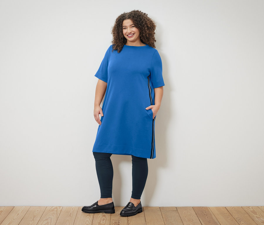 Une femme aux cheveux bouclés porte une robe bleue avec fermeture éclair et des leggings, posant devant un mur.