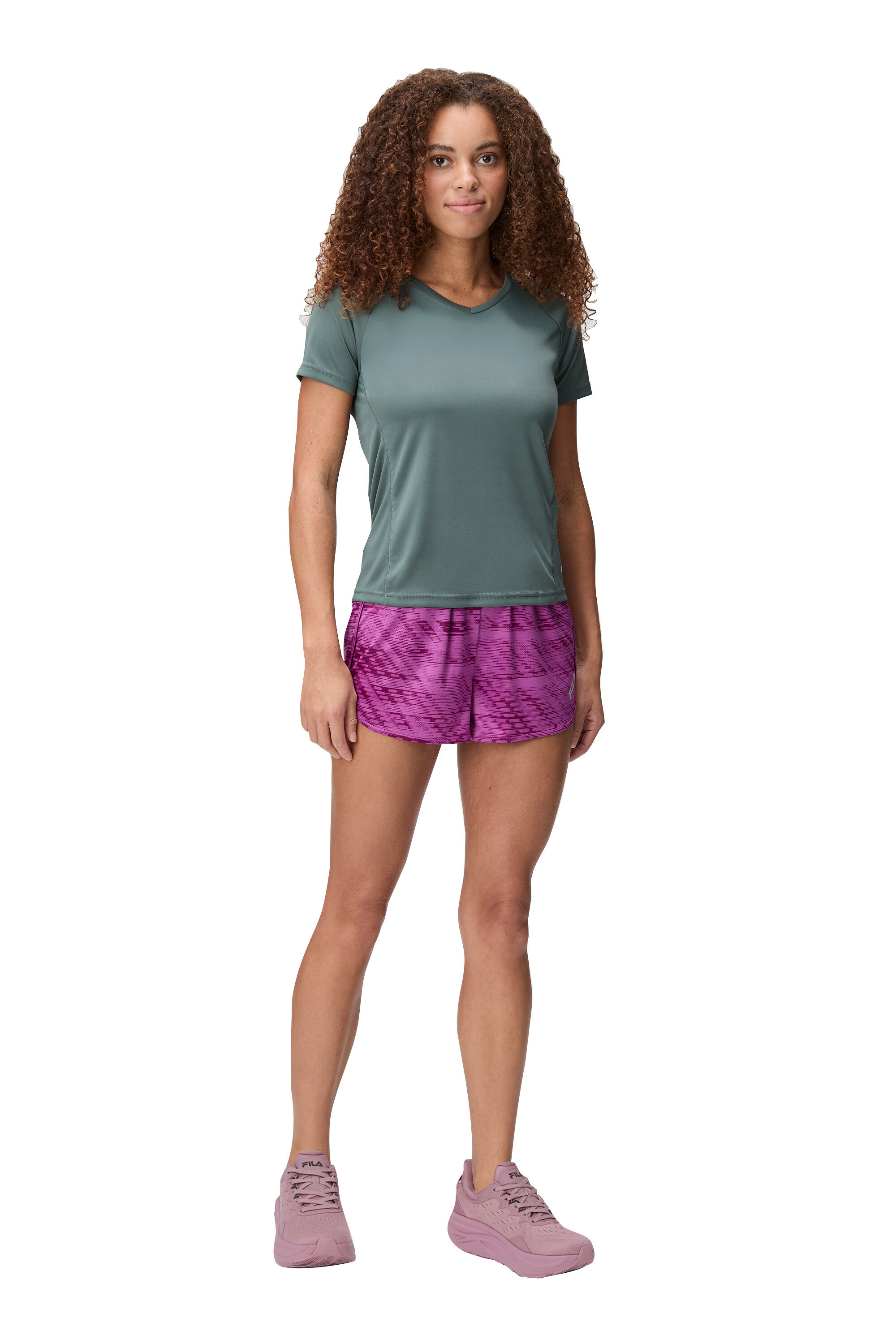 Femme debout sur fond blanc portant un t-shirt vert, un short rose et des chaussures roses.