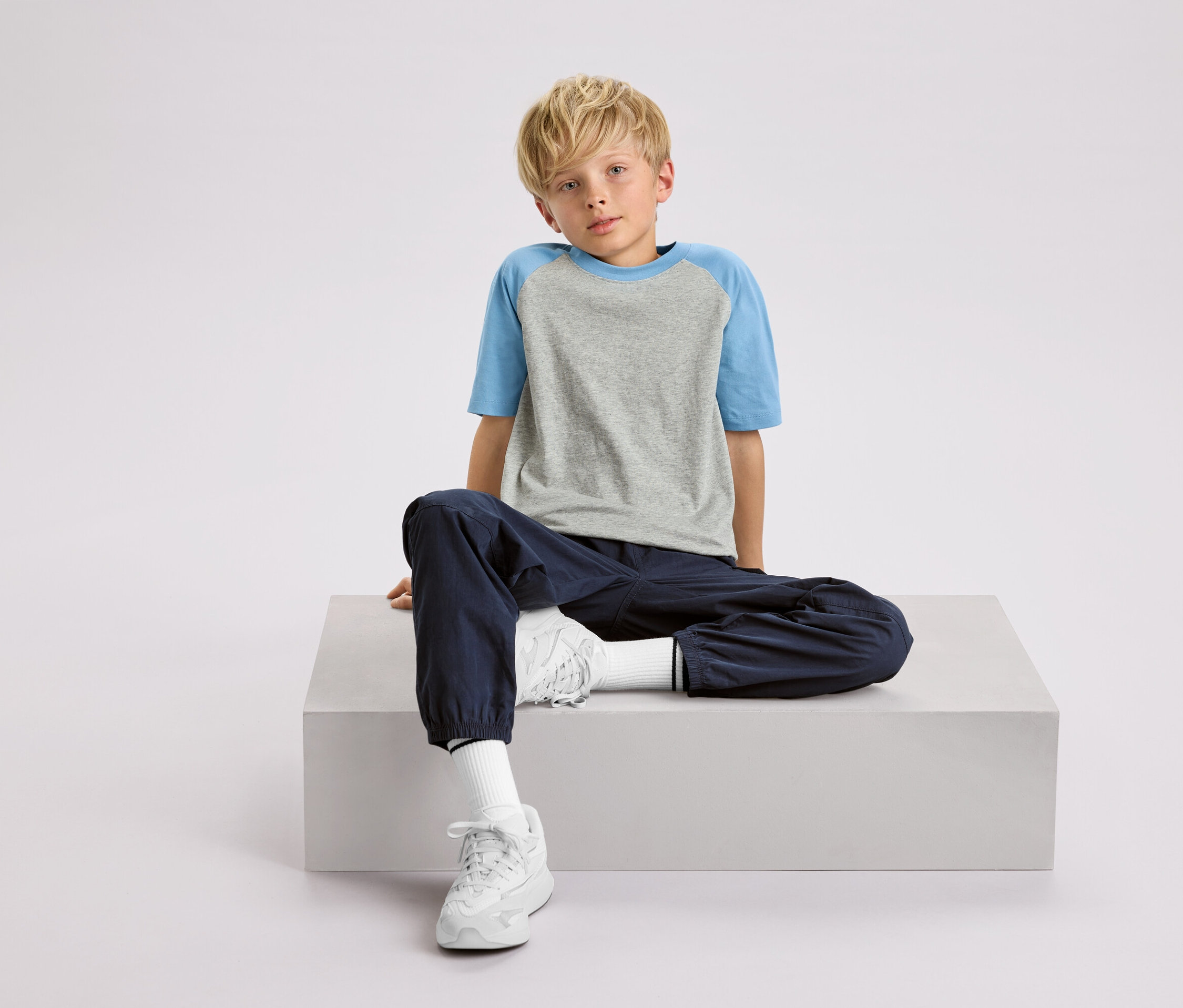 Un garçon blond est assis sur un cube blanc, portant un t-shirt gris et bleu, un pantalon bleu marine et des baskets blanches.