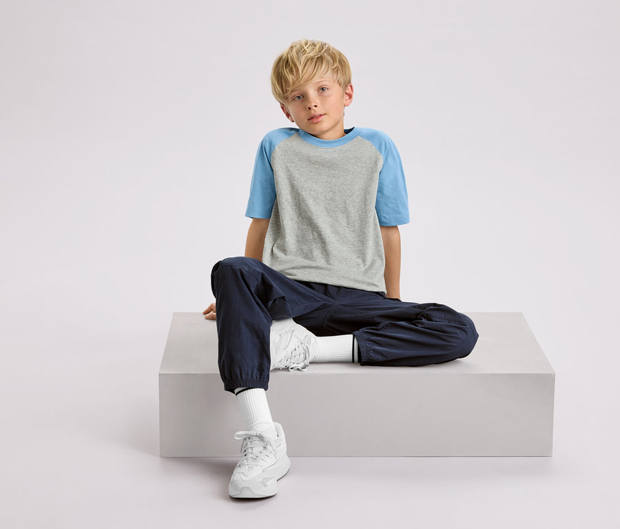 Un garçon blond est assis sur un cube blanc, portant un t-shirt gris et bleu, un pantalon bleu marine et des baskets blanches.