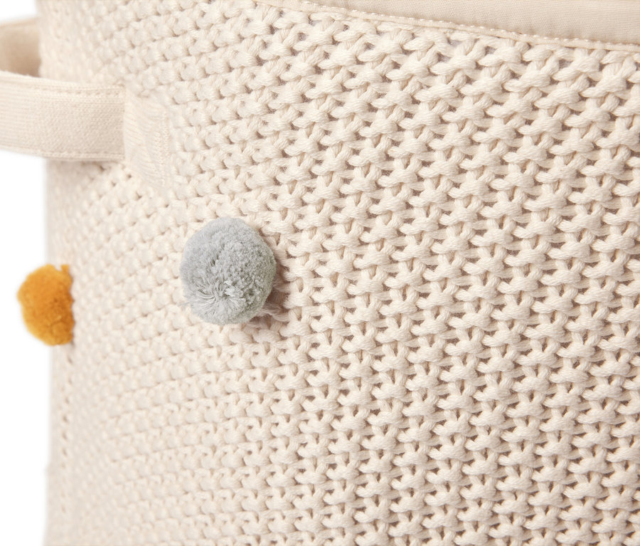 Gros plan sur un panier de rangement en tricot crème avec une poignée et des pompons jaunes et gris.